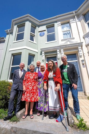 Six people - three women and three men standing on the front path of a terraced house. They are all smiling. Behind them is the front of the house with a new BluePlaque attached to the outside wall of the first floor, between two windows.