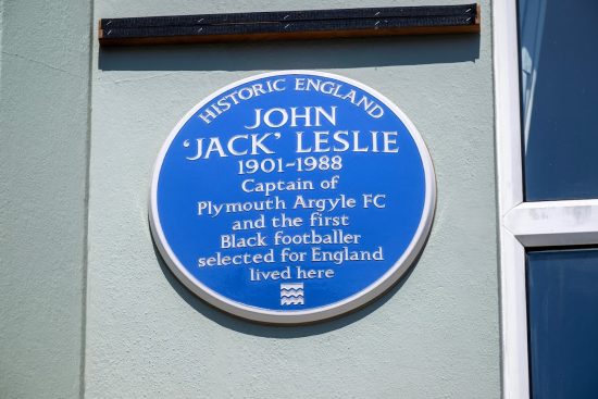 A close-up colour photo of a round blue plaque mounted on a grey wall. The text on the plaque reads: 'Historic England- John 'Jack' Leslie 1901-1988 - Captain of Plymouth Argyle FC and the first Black footballer selected for England lived here' At the bottom is Historic England's logo.