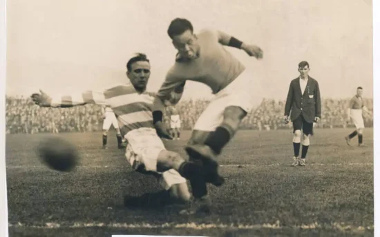 A monochrome action photo of a football match from the 1920s. One player, a white man in a hooped shirt and pale shorts, is attempting to tackle a second player, a black man in a dark shirt and pale shorts. The ball is blurred as it is moving so fast. In the background, the referee and another player are watching the action, and there is a large crowd in the stands in the distance.
