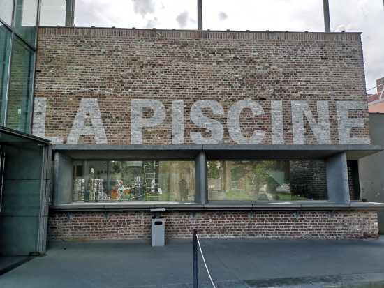 The outside of a brick and glass building. The words 'La Piscine' are painted in grey letters on the brick.