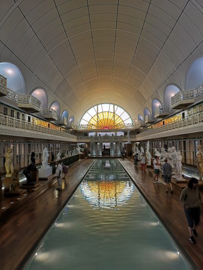 The inside of an art gallery built around a swimming pool. The pool, filled with water, runs through the centre of the gallery. On the poolside are statues of human, and visitors walking around. At the end of the pool is a yellow and orange stained glass window depicting the sun.