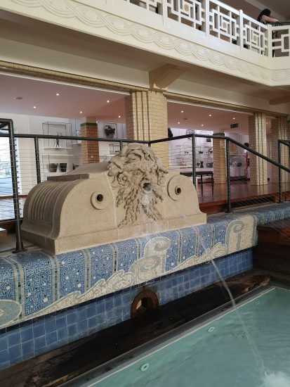 A water fountain in the shape of a male human head, made of a grey stone. It is resting on a blue tiled wall. A jet of water is shooting from the man's mouth into a pool of water.