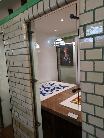 A small room made of off-white ceramic bricks with a glass window. Behind the window is a display case showing two designs on paper, one in blue and white and one in red and yellow. A portrait of a woman hanging on the wall at the back.