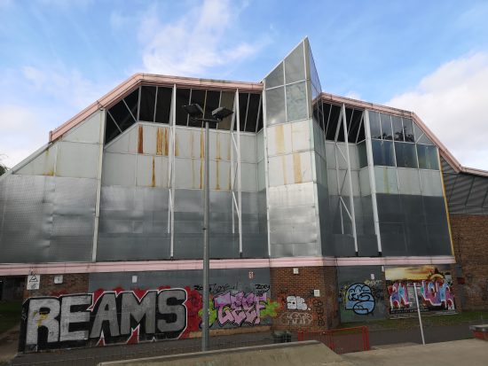 An exterior view of a building. The lower level is made of brick, and the upper level are plastic and steel. All of the windows are covered by metal shutters, and there is graffiti on the brick level.