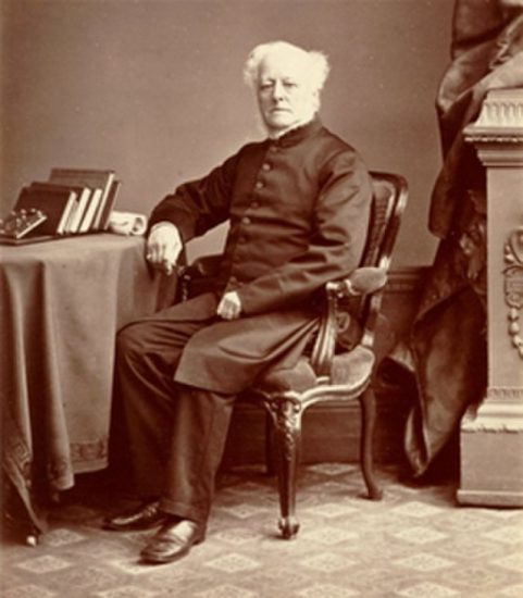 A sepia/monotone photograph from the 1870s of a white man sitting at a desk and looking at the camera. He is dressed in a formal dark suit. There are some books on the desk.